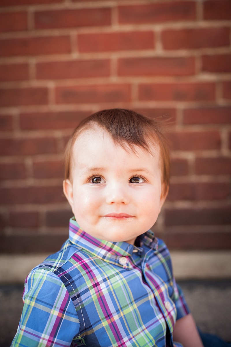 Adorable one year old boy in plaid next to brick wall